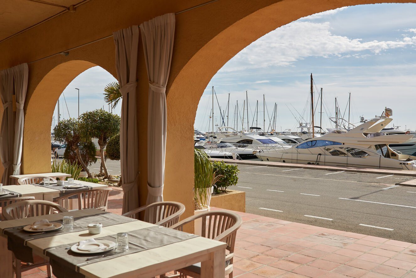 La terrasse sous arcades de Saltea, tables dressées et bateaux de la marina en arrière-plan